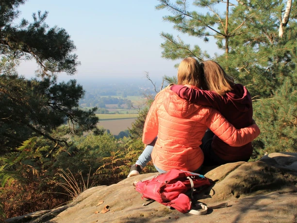 Ausblick von den Dörenther Klippen Zwei Personen sitzen auf einem Felsen, umgeben von Kiefern, und blicken auf eine weite Landschaft.