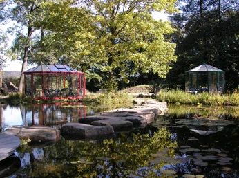 Teich im NaturaGart-Park Steinpfad führt über ruhigen Teich mit Seerosen, flankiert von zwei Pavillons und üppigen Bäumen.