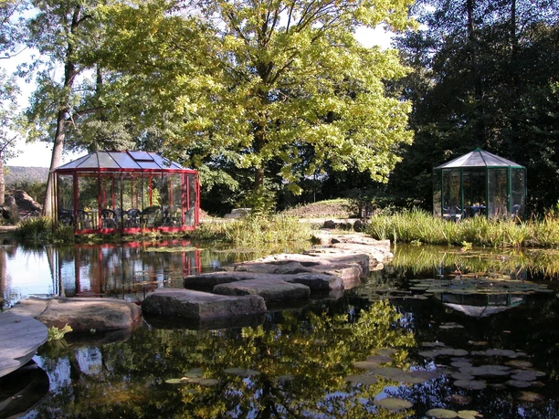 Teich im NaturaGart-Park Steinpfad führt über ruhigen Teich mit Seerosen, flankiert von zwei Pavillons und üppigen Bäumen.
