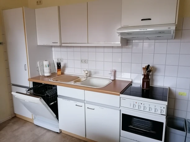 Küche 1 Helle Küche mit weißen Schränken, Holzarbeitsplatte, Spüle, Herd und geöffneter Spülmaschine.