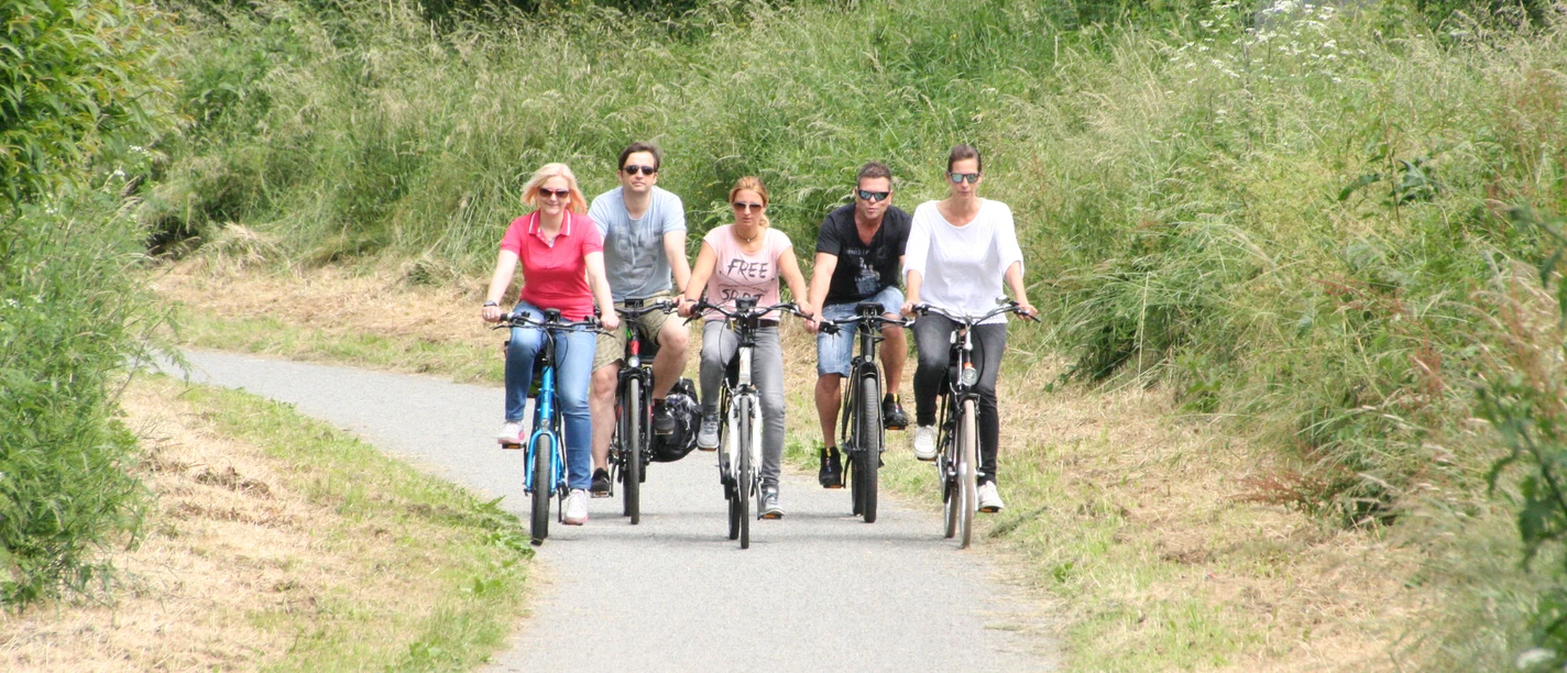 Radler Mittelweser Fünf Menschen radeln entspannt auf einem schmalen asphaltierten Weg inmitten dichter grüner Vegetation.