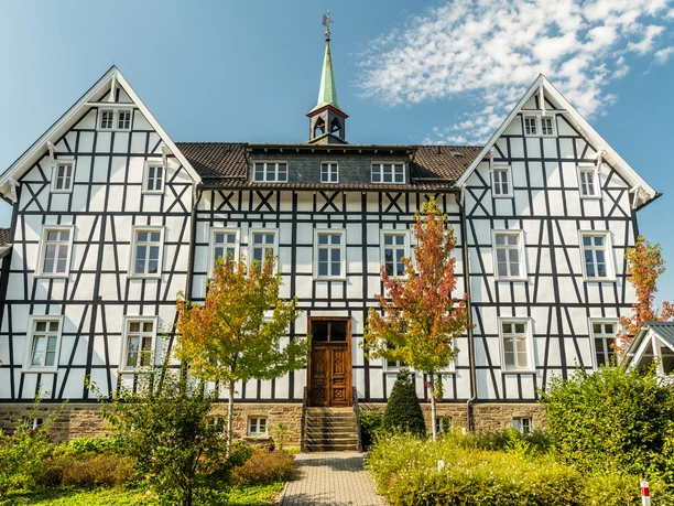 Altes Kloster Much Fachwerkhaus mit grünem Turm und blauen Himmel im Hintergrund; flankiert von bunten Herbstbäumen.