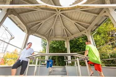 Kneipp-Vitalpark Eine Frau und ein Mann entspannen sich und unterhalten sich in einem hölzernen Pavillon im Freien.