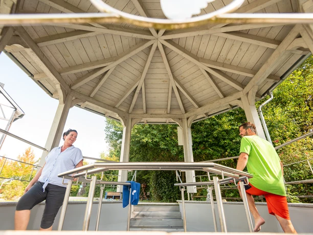 Kneipp-Vitalpark Eine Frau und ein Mann entspannen sich und unterhalten sich in einem hölzernen Pavillon im Freien.