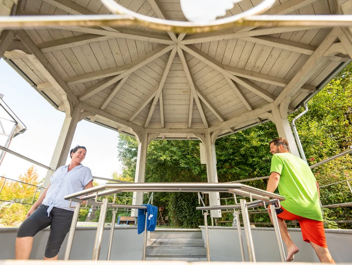 Kneipp-Vitalpark Eine Frau und ein Mann entspannen sich und unterhalten sich in einem hölzernen Pavillon im Freien.