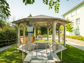 Kneipp-Vitalpark Ein barrierefreier Pavillon aus Holz mit rampe am Rande eines grünen Gartens bei sonnigem Wetter.