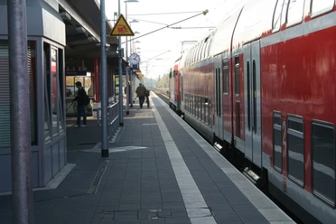 Bahnsteig Achim mit wartenden Passagieren und rotem Zug, eingefasst von Überdachung und Schienen.