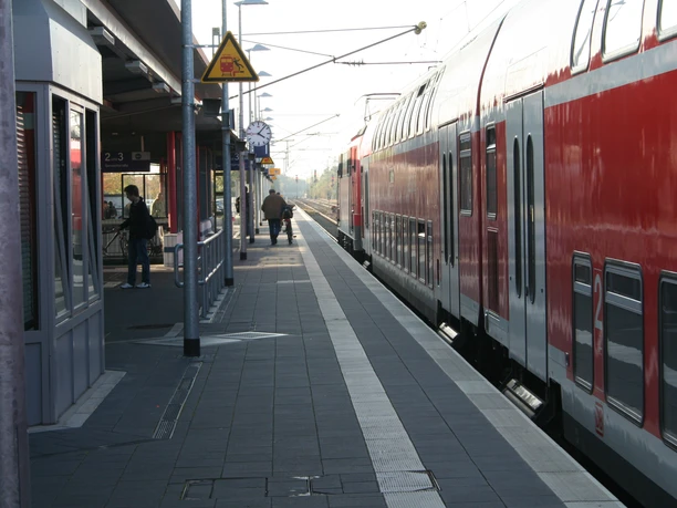Bahnsteig Achim mit wartenden Passagieren und rotem Zug, eingefasst von Überdachung und Schienen.
