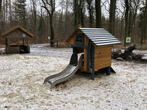 Kleines Holzspielhaus mit silberner Rutsche in einem verschneiten Waldstück mit angrenzendem Unterstand.
