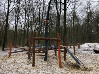 Moderner Waldspielplatz mit Klettergerüst, Rutsche und Seilstrukturen in winterlichem Waldambiente.
