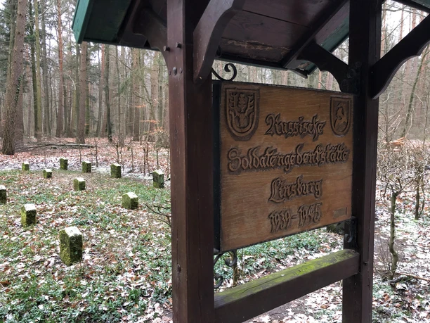 Holztafel mit Inschrift "Russische Soldatengräber 1945–1945" vor bescheidenem Waldfriedhof.