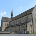 Mittelalterliche Klosterkirche aus Stein mit rotem Turm vor blauem Himmel, umgeben von Gärten.
