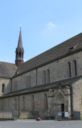 Mittelalterliche Klosterkirche aus Stein mit rotem Turm vor blauem Himmel, umgeben von Gärten.