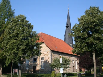Steingebäude mit rotem Ziegeldach und hohem Turm; von grünen Bäumen umgeben, blauer Himmel.