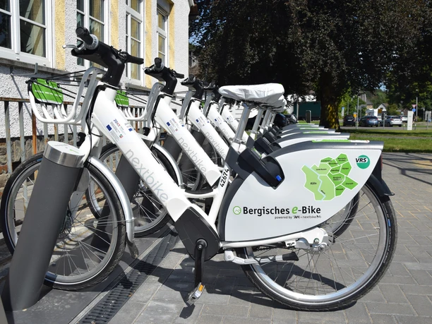 Bergisches E-Bike Reihe von weißen E-Bikes zur Miete, aufgereiht an einer Dockingstation im Freien.