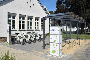 Bergisches E-Bike Fahrradverleihstation mit Schutzdach, mehreren Fahrrädern und Bedienungssäule auf gepflastertem Boden.
