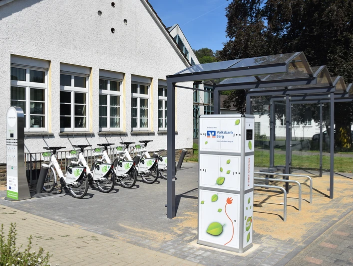 Bergisches E-Bike Fahrradverleihstation mit Schutzdach, mehreren Fahrrädern und Bedienungssäule auf gepflastertem Boden.