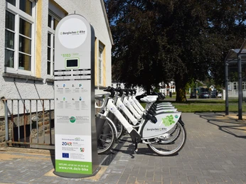 Bergisches E-Bike Fahrradverleihstation mit weißen E-Bikes neben einem Gebäude und grünen Bäumen im Hintergrund.
