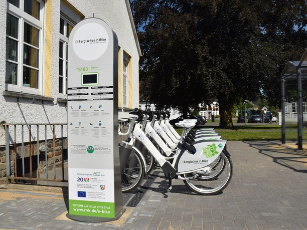 Bergisches E-Bike Fahrradverleihstation mit weißen E-Bikes neben einem Gebäude und grünen Bäumen im Hintergrund.