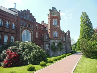 Brauerei Strate Historische Backstein-Brauerei mit Türmchen, großen Fenstern, umgeben von gepflegtem Grün und Rosen.