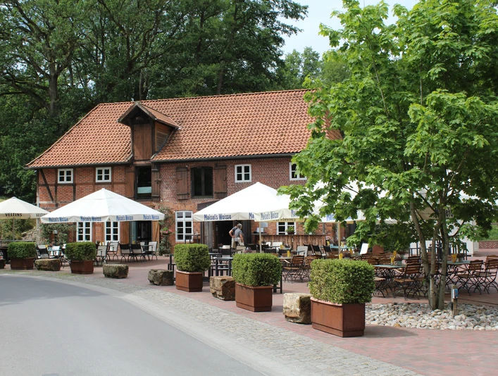 Historische Backsteinmühle mit Biergarten, Holztischen und weißen Sonnenschirmen unter blühendem Baum.