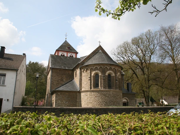 Ostansicht St. Pankratius Odenthal Romanische Kirche mit halbrunden Apsiden, Turm mit Zinnen, umgeben von Bäumen und einem Steinzaun.
