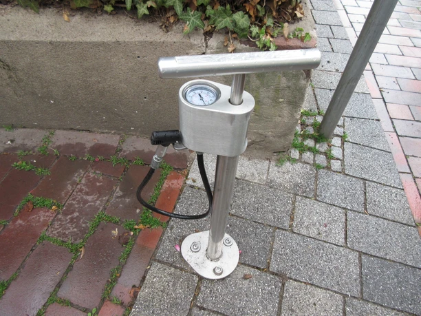 Lufttankstelle aus Metall mit Manometer und Schlauch auf gepflastertem Gehweg neben einem Betonrand.
