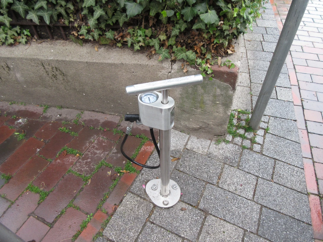 Luftstation mit Handpumpe für Fahrräder auf einem gepflasterten Bürgersteig, von Pflanzen umgeben.