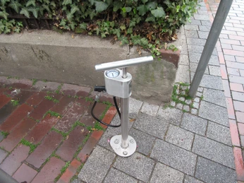 Luftstation mit Handpumpe für Fahrräder auf einem gepflasterten Bürgersteig, von Pflanzen umgeben.