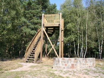 Januarsberg Turm Ein hölzerner Aussichtsturm mit steinerner Umgrenzung, umgeben von Bäumen und blauem Himmel.