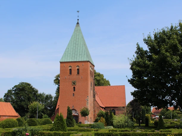 Riede church with red brick tower, green pointed roof, surrounded by trees and well-tended garden.