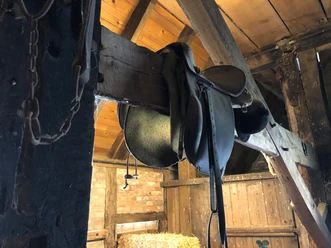 Antiker Reitsattel hängt in einem rustikalen Holzstall mit Strohballen und alten Holzbalken.