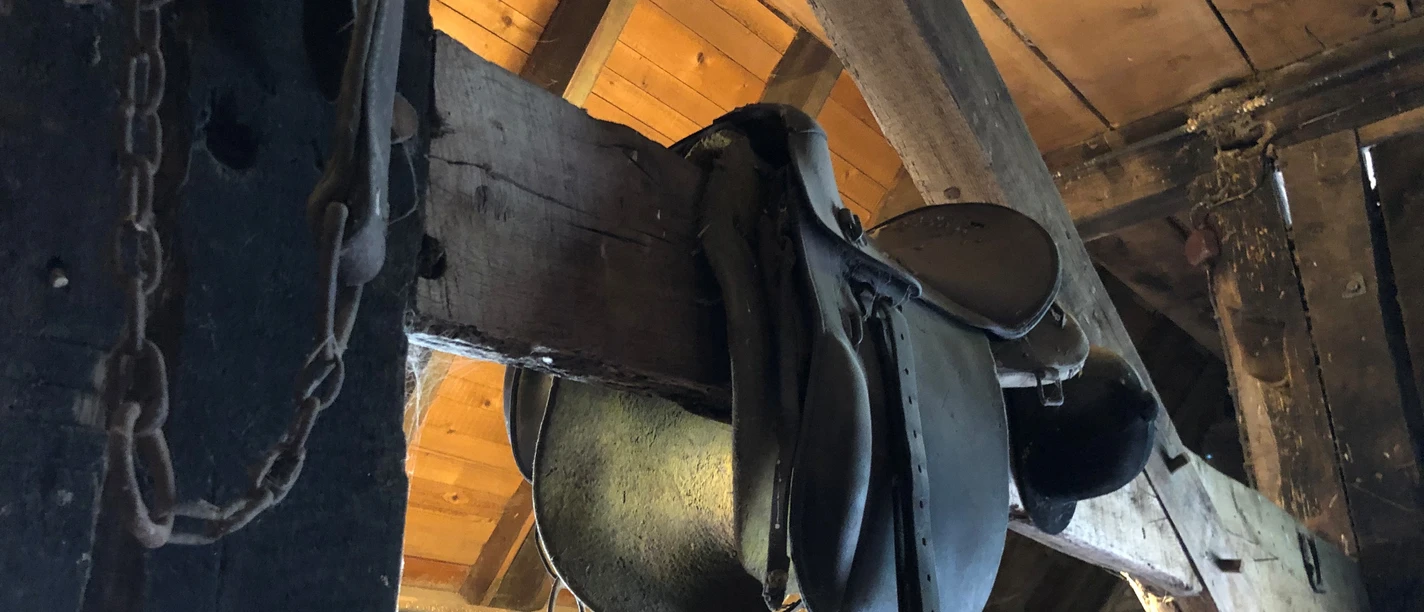 Antiker Reitsattel hängt in einem rustikalen Holzstall mit Strohballen und alten Holzbalken.