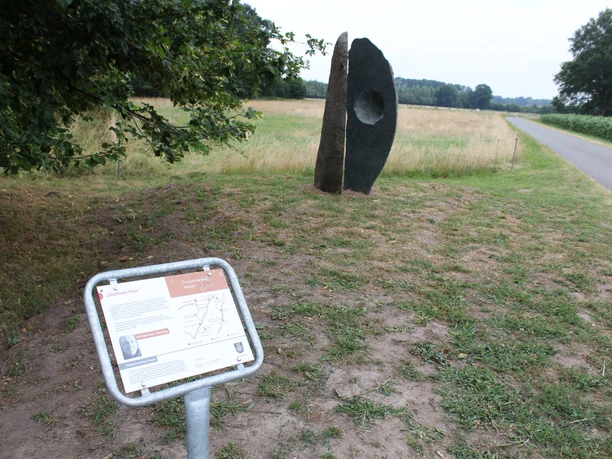 Sculptuur van donkere steen met een ovale opening, staat aan de rand van het veld naast een informatiebord.