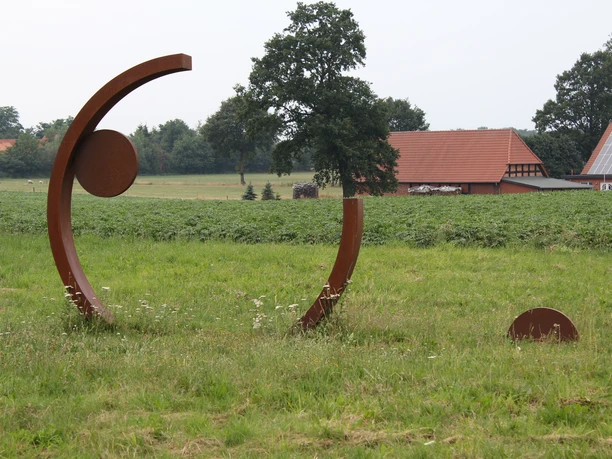 Een moderne metalen sculptuur staat op een groene weide, omringd door bomen en landelijke architectuur.