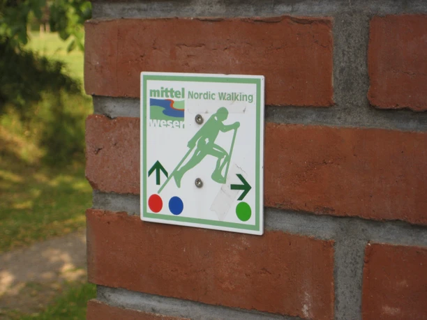Nordic Walking Thedinghausen Wegweiser für Nordic Walking, befestigt auf einer Backsteinmauer, umgeben von grünem Laub.