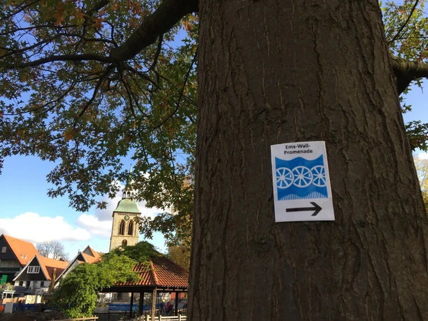 Qualitätsweg Ems-Wall-Promenade: Markierung am Emssee Schild der Ems-Wall-Promenade an einem Baum, im Hintergrund eine Kirchturmspitze und bunte Herbstbäume.