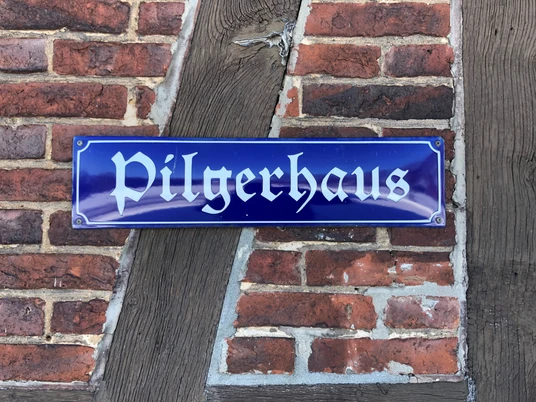 Blaues Metallschild mit gotischer Aufschrift "Pilgerhaus" an einer rustikalen Backstein-Holz-Fassade befestigt.
