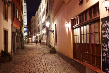 Abendstimmung in einer Altstadtgasse von Köln, belebt von zwei Personen auf Kopfsteinpflaster.