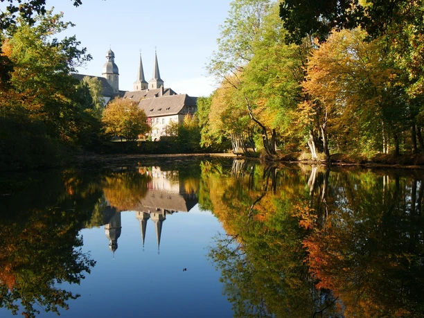 Blick auf die Abtei Marienmünster im Herbst Abtei Marienmünster spiegelt sich im ruhigen See, umgeben von herbstlichen Bäumen in leuchtenden Farben.