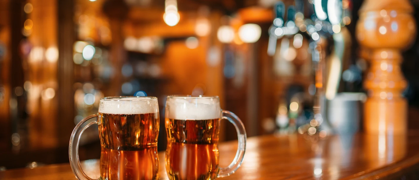 Two beer mugs on wooden bar counter, macro, nobody. Octoberfest symbol or concept. Glasses with golden beverage and foam on the table in pub, closeup view, blur background