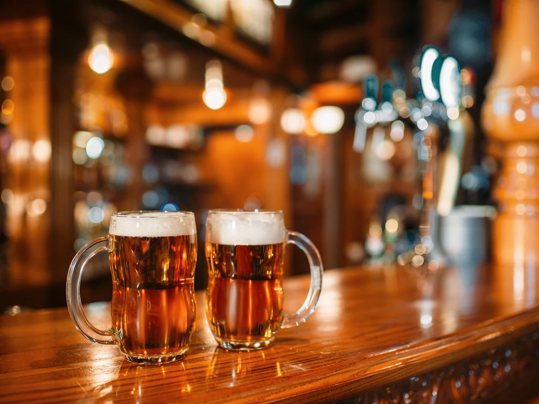 Two beer mugs on wooden bar counter, macro, nobody. Octoberfest symbol or concept. Glasses with golden beverage and foam on the table in pub, closeup view, blur background Two beer mugs on wooden bar counter, macro, nobody. Octoberfest symbol or concept. Glasses with golden beverage and foam on the table in pub, closeup view, blur background