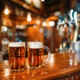 Two beer mugs on wooden bar counter, macro, nobody. Octoberfest symbol or concept. Glasses with golden beverage and foam on the table in pub, closeup view, blur background Two beer mugs on wooden bar counter, macro, nobody. Octoberfest symbol or concept. Glasses with golden beverage and foam on the table in pub, closeup view, blur background
