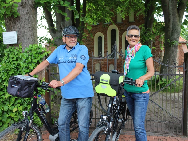Radler Achim Zwei Radfahrer mit Helmen vor einem historischen Backsteingebäude, umgeben von grünen Bäumen.