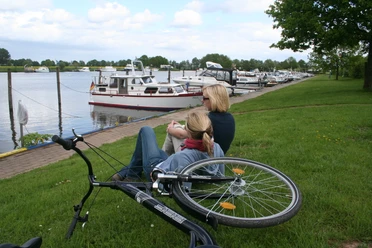 Two women sit on the bank of a river with a view of anchored boats, a bicycle lying next to them.