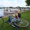 Zwei Frauen sitzen am Ufer eines Flusses mit Blick auf ankernde Boote, ein Fahrrad liegt daneben.