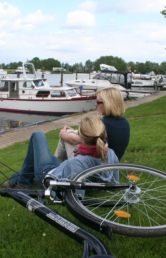 Mittelweser-Region-Radfahren Zwei Frauen sitzen am Ufer eines Flusses mit Blick auf ankernde Boote, ein Fahrrad liegt daneben.