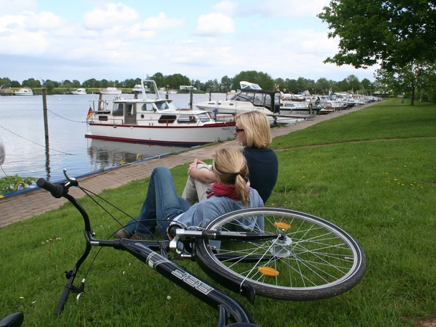 Mittelweser-Region-Radfahren Zwei Frauen sitzen am Ufer eines Flusses mit Blick auf ankernde Boote, ein Fahrrad liegt daneben.