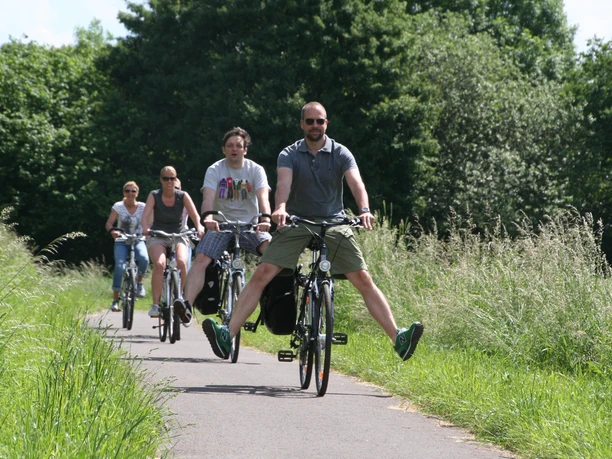 Radfahren Mittelweser Eine Gruppe von Menschen radelt entspannt auf einem schmalen Weg durch grüne, üppige Natur.