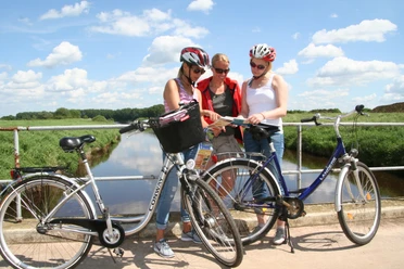 Radfahren Mittelweser Drei Radfahrer mit Helmen stehen auf einer Brücke über einem Fluss und betrachten gemeinsam eine Karte.